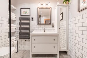 Une salle de bain moderne avec des murs recouverts de carreaux blancs, un lavabo blanc avec un miroir au-dessus, et un porte-serviettes où sont rangées des serviettes pliées avec soin.