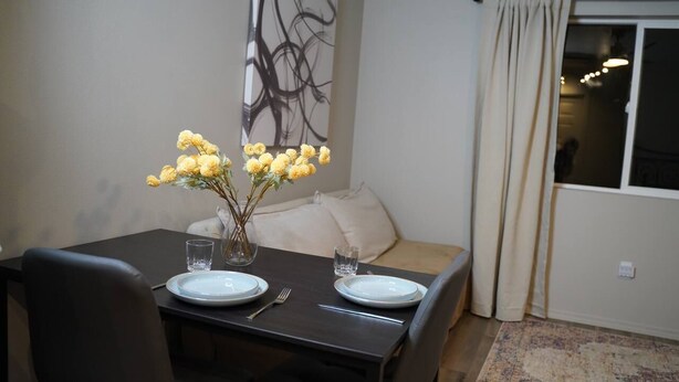 A dining area with a table set for two, a vase with yellow flowers, and a modern abstract painting on the wall.