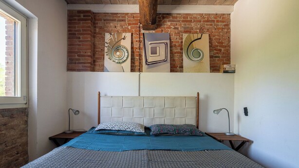 Un dormitorio con pared de ladrillo, una cama con ropa de cama estampada, dos lámparas de noche y arte abstracto en la pared.