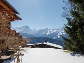 A wooden cabin with a snow-covered roof, a clear view of snow-capped mountains, and a pine tree to the right. - Chaperon Rouge B5 by Interhome (Villars)