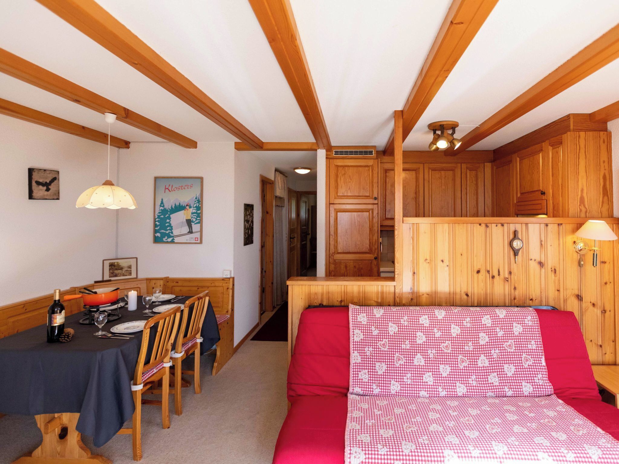 A cozy room with a dining area, a bed with a red and white checkered bedspread, wooden ceiling beams, and a wooden cabinet.