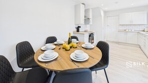 A modern dining area with a round table set for six, black upholstered chairs, and a kitchen in the background.