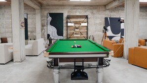 Une table de billard dans une pièce dont le plafond et les murs sont en béton, entourée de meubles blancs et orange.