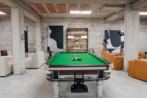 A pool table in a room with a concrete ceiling and walls, surrounded by white and orange furniture.