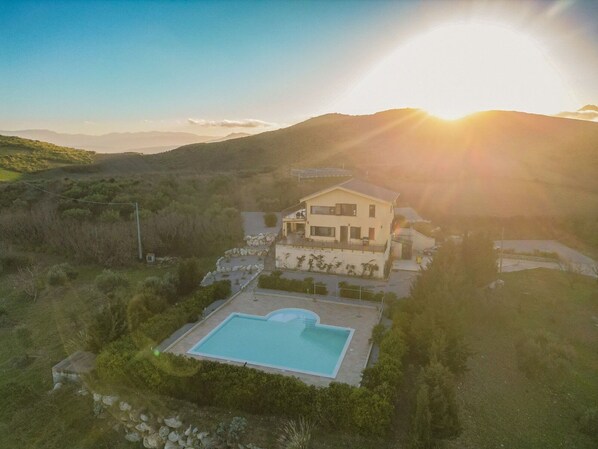 A two-story house with a swimming pool, surrounded by hills and a clear sky.