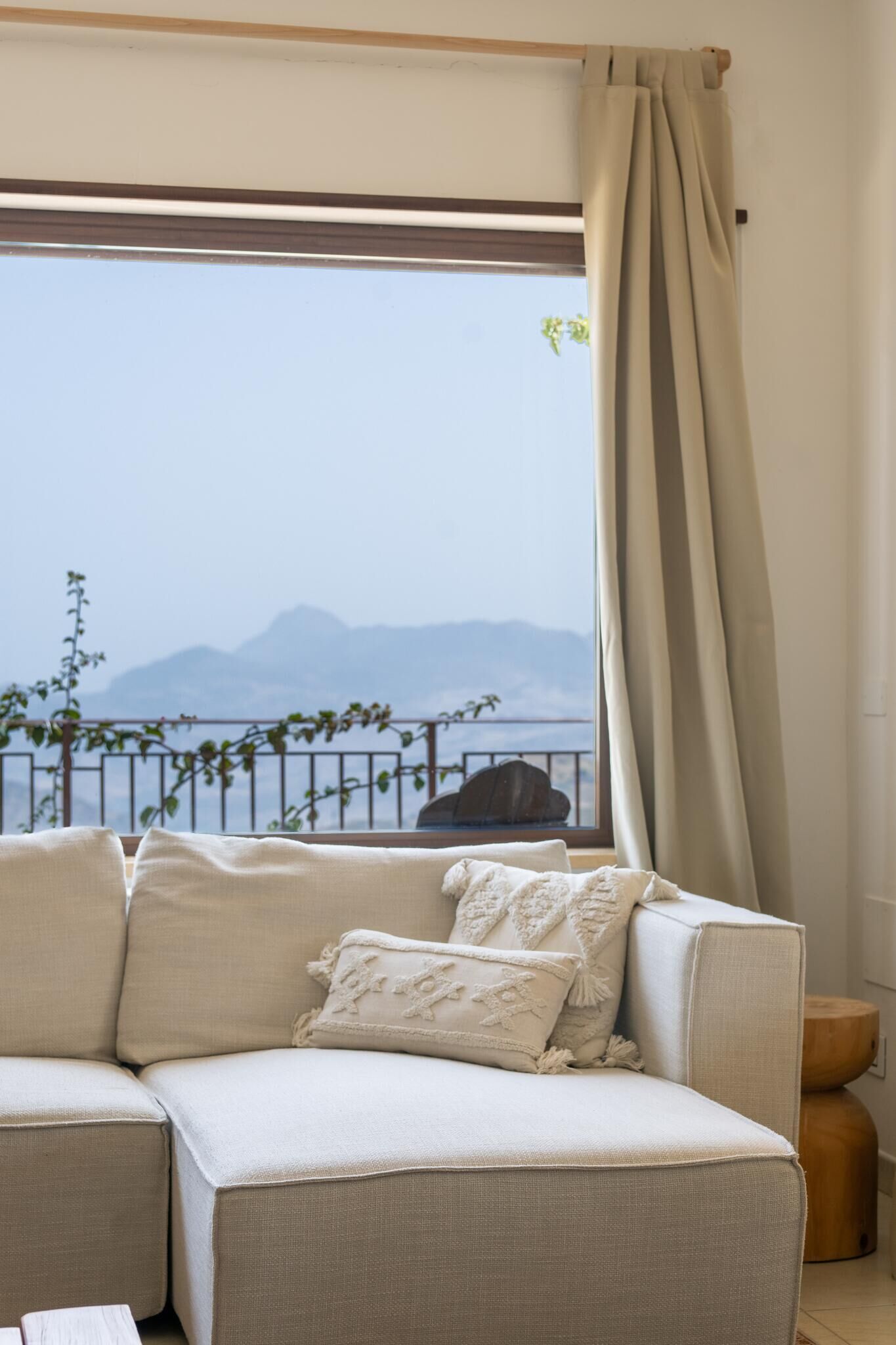 A room with a beige sofa, a wooden side table, and a view of mountains through a window with a curtain.