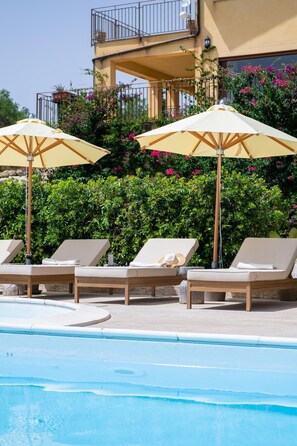Ein Poolbereich mit Liegestühlen und Sonnenschirmen, ein klares, blaues Schwimmbecken und ein Gebäude mit Balkonen und blühenden Pflanzen im Hintergrund.
