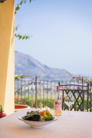 Ein Tisch mit einer Gemüseschale, einer Weinflasche und einem Glas auf einem Balkon mit Bergblick.