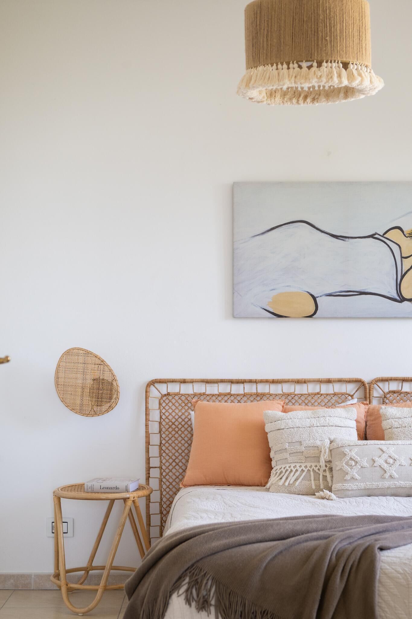 Un dormitorio con cama, cabecera tejida, lámpara colgante, un cuadro en la pared y una pequeña mesa de mimbre.