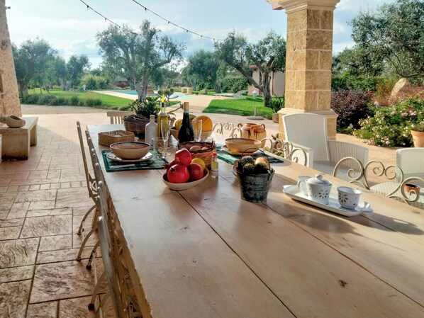 An outdoor dining area with a long wooden table set with various dishes, a potted plant, and a view of a garden and pool.
