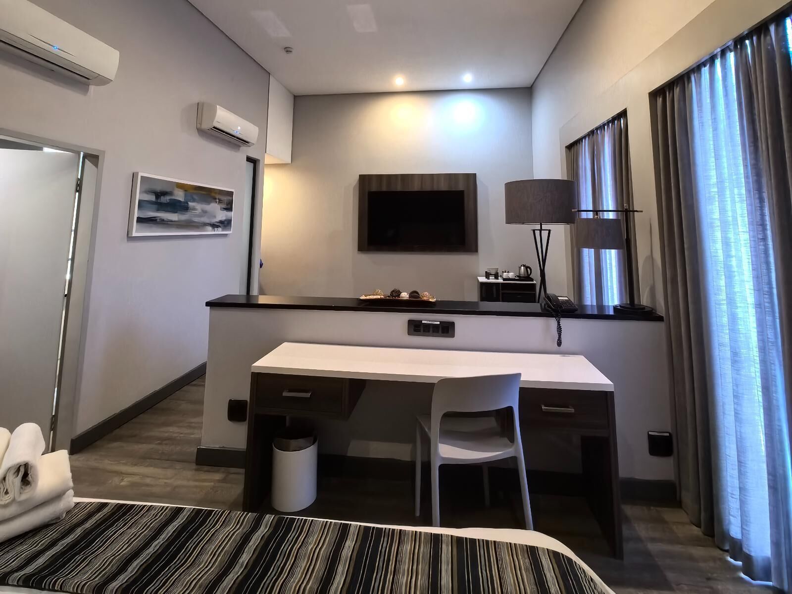 A modern hotel room with a bed, a desk, a chair, a TV, a lamp, and a window with curtains.