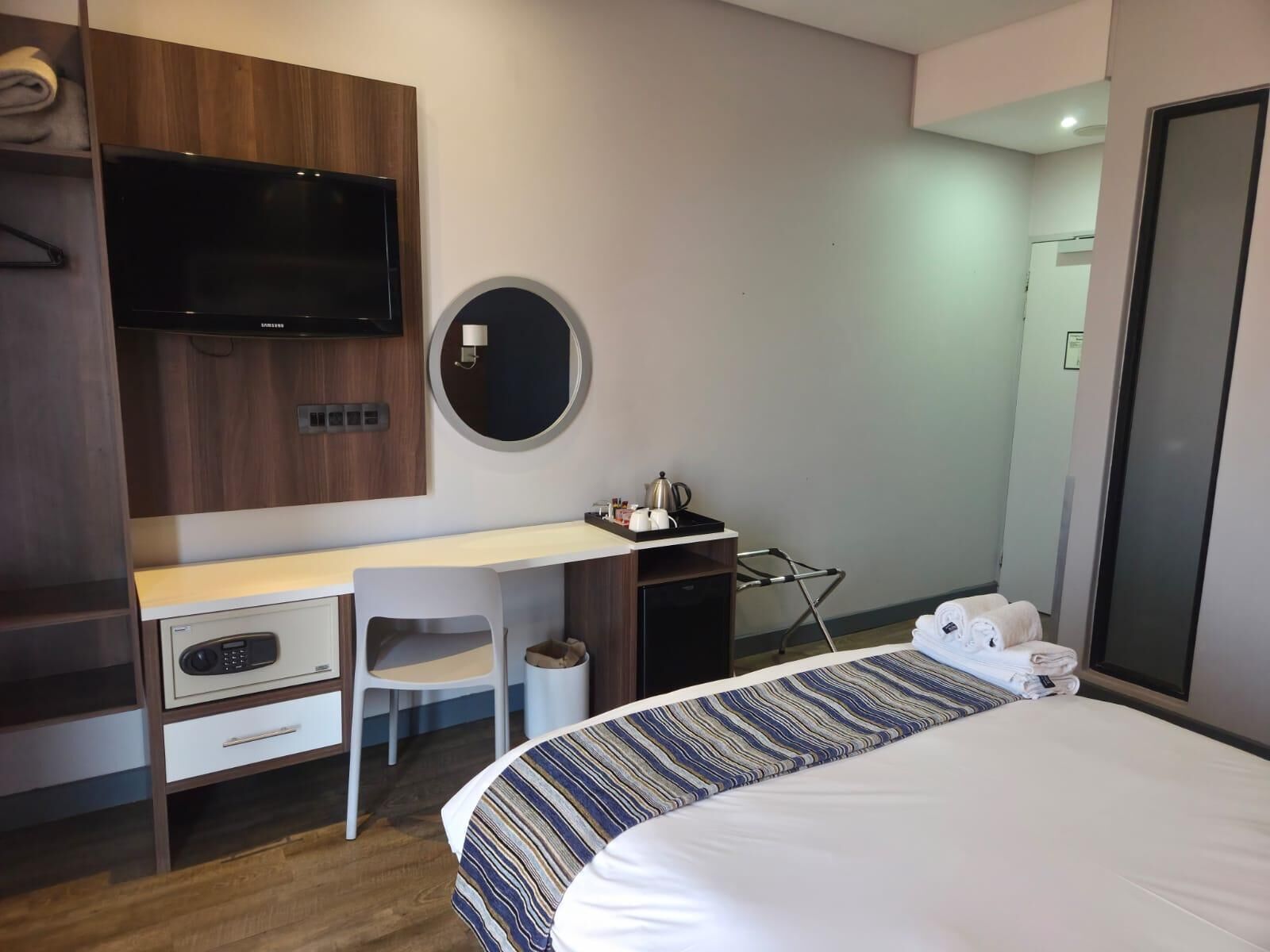 A hotel room with a bed, a desk with a chair, a TV mounted on the wall, a round mirror, and a small table with a kettle and cups.