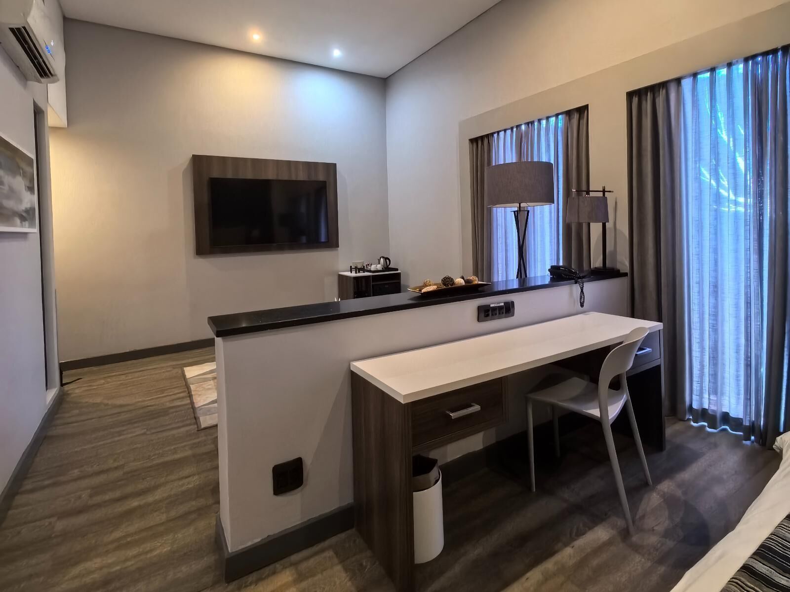 A modern hotel room with a flat-screen TV mounted on the wall, a desk with a chair, a small table with a lamp, and a window with curtains.
