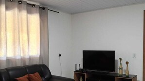 Ein Wohnzimmer mit einem schwarzen Ledersofa, einem Flachbildfernseher auf einem Holzbodenständer, einer kleinen Holztrennwand mit Dekorationen und einem beigen Teppich auf dem gefliesten Boden.