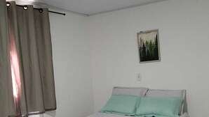 Ein Zimmer mit einem Einzelbett, einer schlichten weißen Wand, einem Gemälde und einem Vorhang.