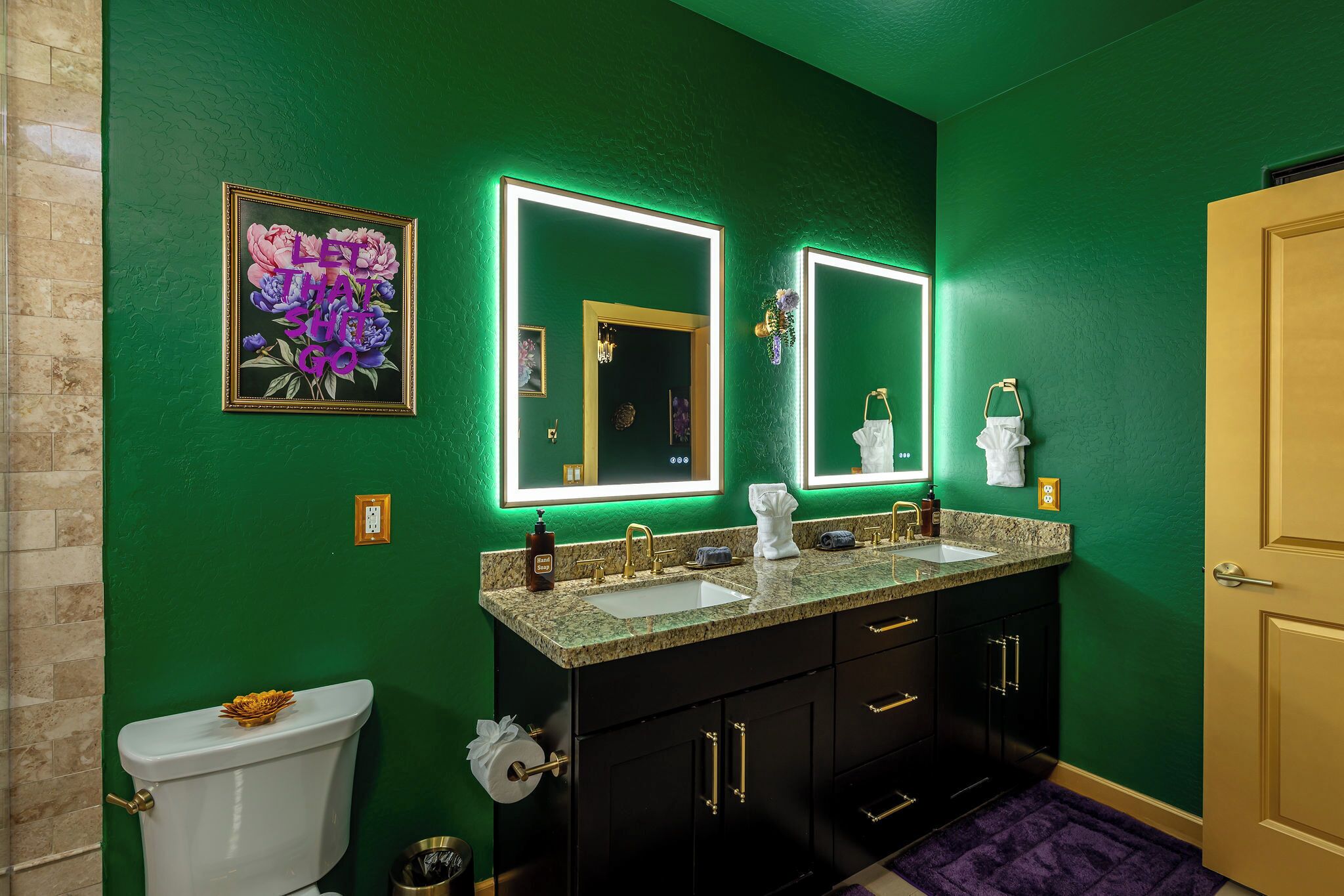 A bathroom with green walls, a white toilet, a double sink vanity, and a framed floral artwork.