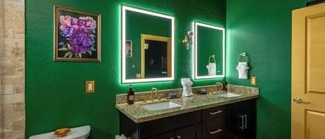A bathroom with green walls, a white toilet, a double sink vanity, and a framed floral artwork.
