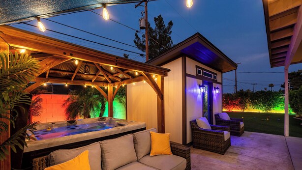 Ein Außenbereich mit Whirlpool, Holzpergola, Liegestühlen und einem kleinen Gebäude mit farbigem Licht.