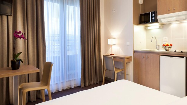 Ein Hotelzimmer mit Bett, Schreibtisch, Stuhl, einer kleinen Küchenzeile und einem Fenster mit Vorhängen.