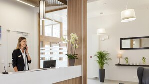 Ein Empfangsbereich mit Rezeptionistin, Empfangstresen, einer Zimmerpflanze, einem Sessel und einem großen Spiegel an der Wand.