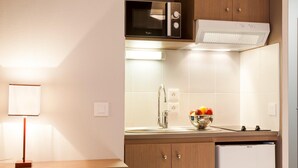 Eine kompakte Küchenzeile mit Mikrowelle, Backofen und kleinem Kühlschrank. Eine Spüle mit Obstschale steht auf der Arbeitsfläche, und ein Essbereich mit Tisch und Stühlen ist vorhanden.
