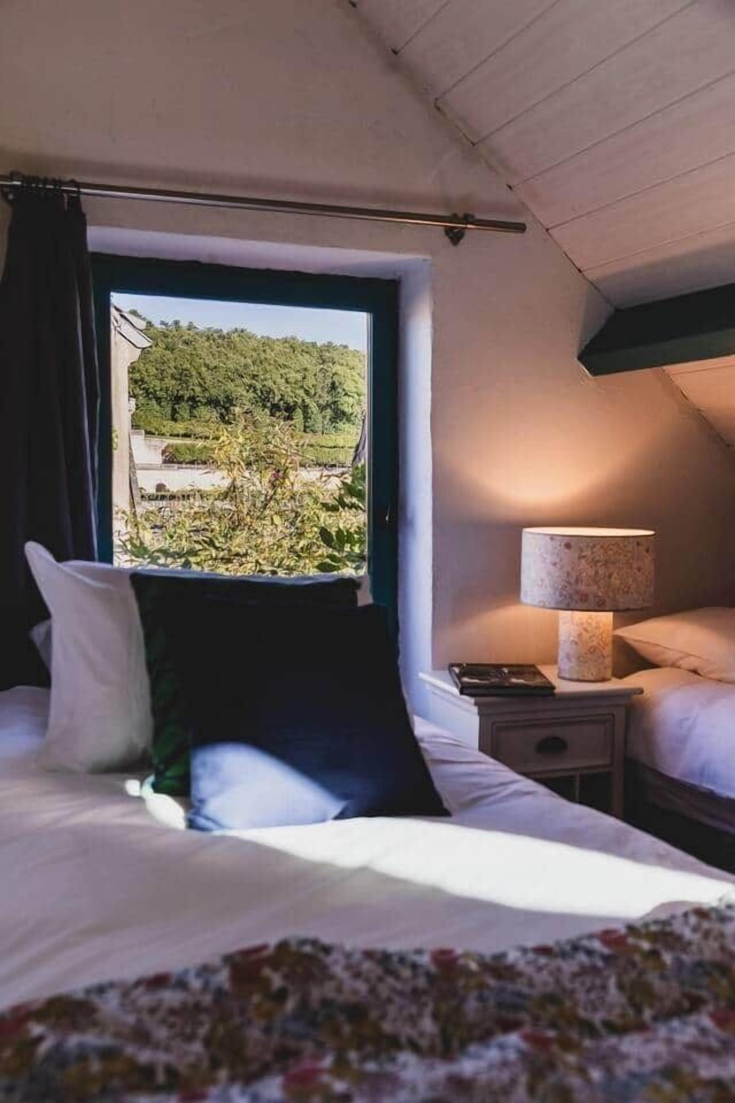 Un dormitorio con cama, mesita de noche, lámpara y ventana con vista a follaje.