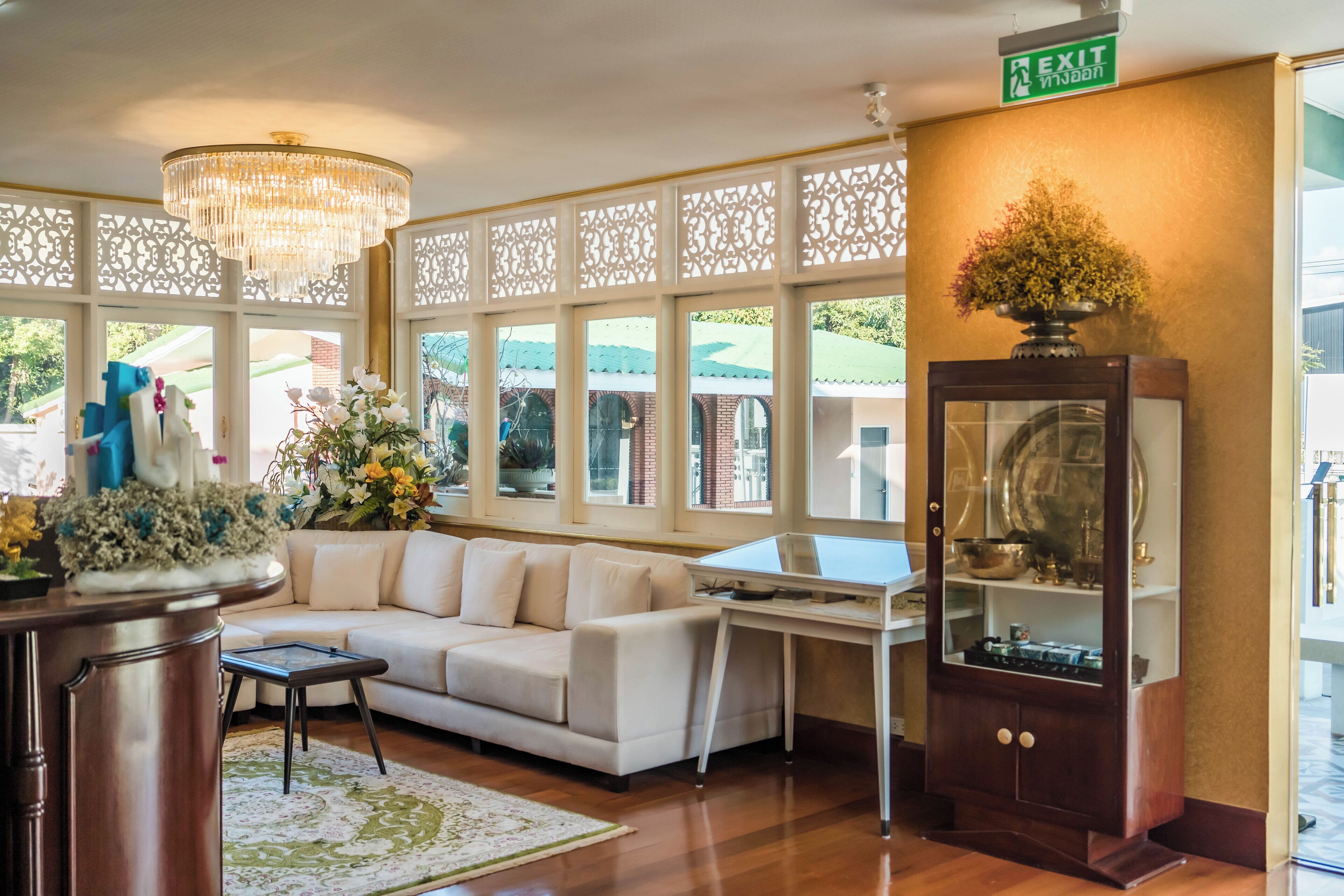 A well-lit living area with a chandelier, a white sofa, a wooden coffee table, and a desk with a computer. There is a decorative plant on the desk and a large window with a view of the outside.