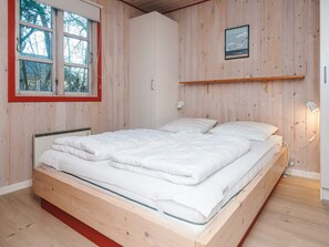 A bedroom with a wooden bed frame, a window with a red frame, and a wall-mounted shelf with a picture.