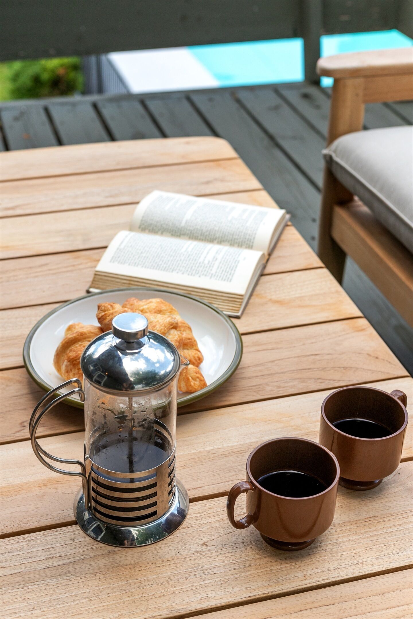 A wooden table with a book, a plate of pastries, a coffee press, and two mugs.