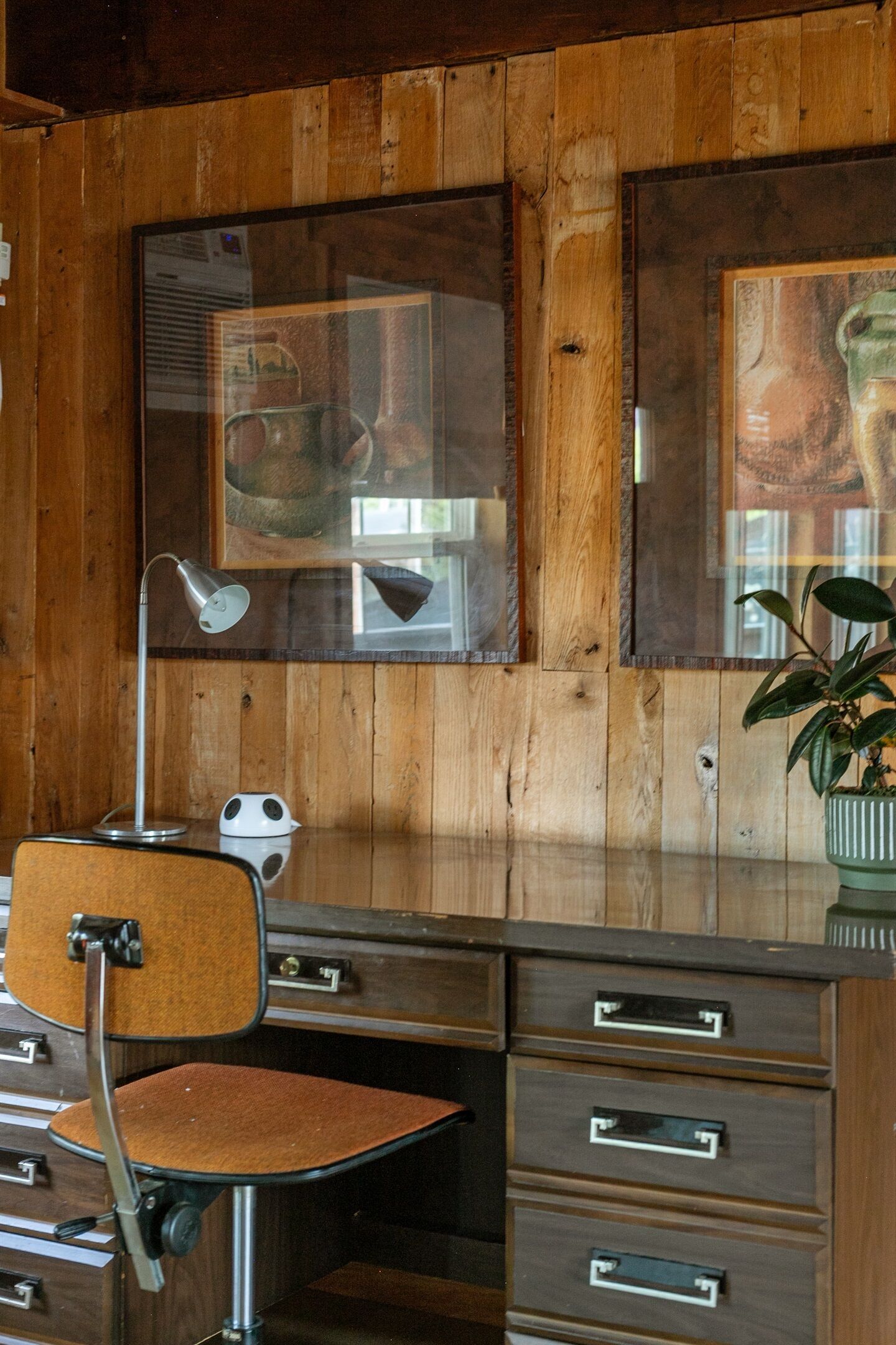 Une pièce aux murs lambrés, avec un bureau, des tiroirs, une chaise, une lampe de bureau et une plante en pot.