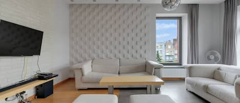 A modern living room with a flat-screen TV, a sectional sofa, a coffee table, and a large window with a city view.