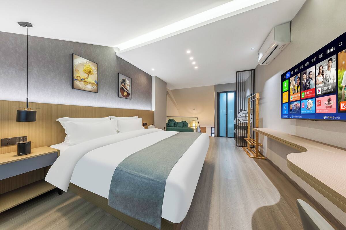A modern hotel room with a large bed, a bedside table, a wall-mounted TV, and a view of the hallway.