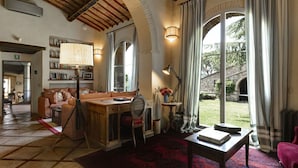 Ein gemütliches Wohnzimmer mit einer Couch, einem Holztisch und einem roten Teppich mit Muster. Ein Bücherregal mit Büchern und einer Lampe steht dort. Das Zimmer hat ein großes Fenster mit Vorhängen und Blick ins Freie.