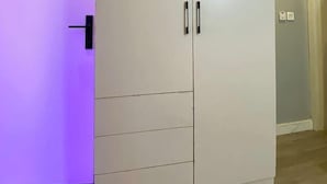 A large white wardrobe with two doors and a handle, located in a room with wooden flooring and a door with purple lighting.