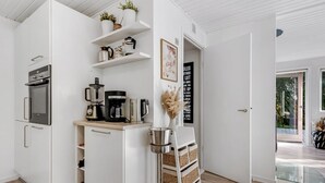 A modern kitchen with white cabinetry, a built-in oven, and a coffee maker. There are shelves with decorative items and a vase with dried flowers.
