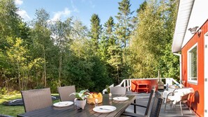 An outdoor dining area with a wooden table and wicker chairs, surrounded by a deck and a red building.