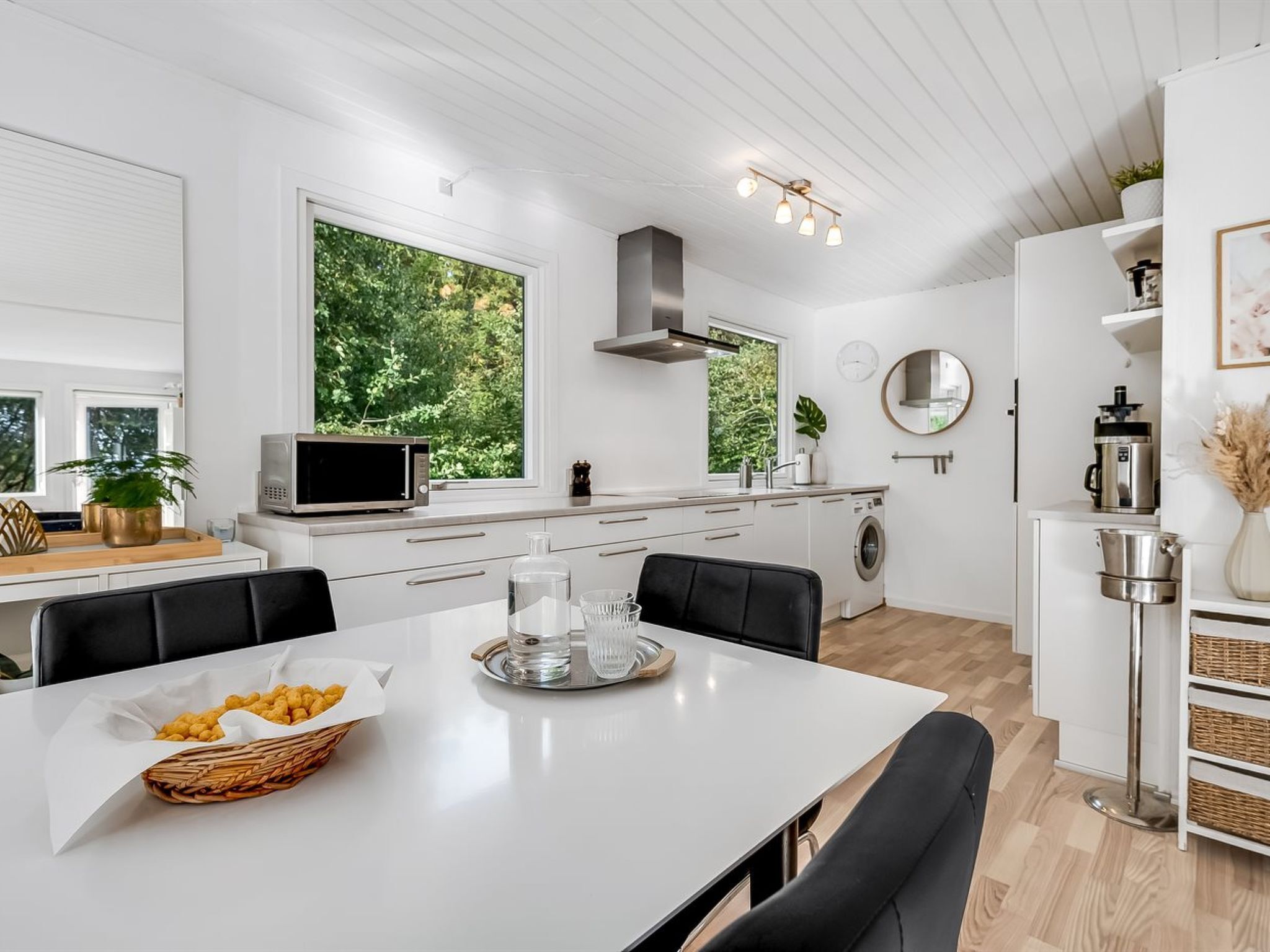 Een moderne keuken met witte kastjes, een eettafel met stoelen, een magnetron, een spoelbak en een groot raam met uitzicht op groen.