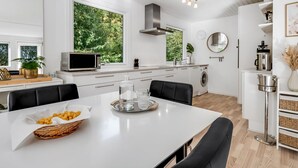 A modern kitchen with white cabinetry, a dining table with chairs, a microwave, a sink, and a large window with a view of greenery.