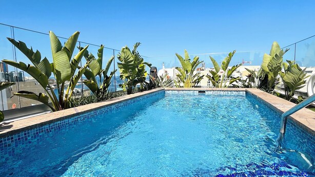 A clear blue pool with mosaic tiles, surrounded by tropical plants and a clear sky. - Sliema Studios (Sliema)