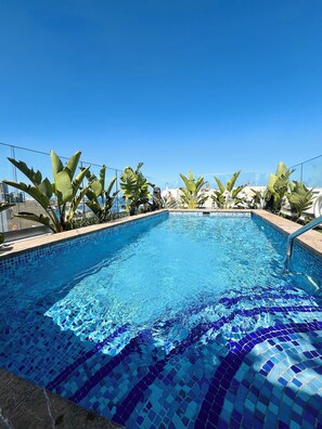 Una piscina azul cristalina con mosaicos, rodeada de plantas tropicales y un cielo despejado.