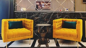A lobby with two yellow armchairs, a marble floor, a small round table, and a TV mounted on a wooden panel wall. - Sliema Studios (Sliema)