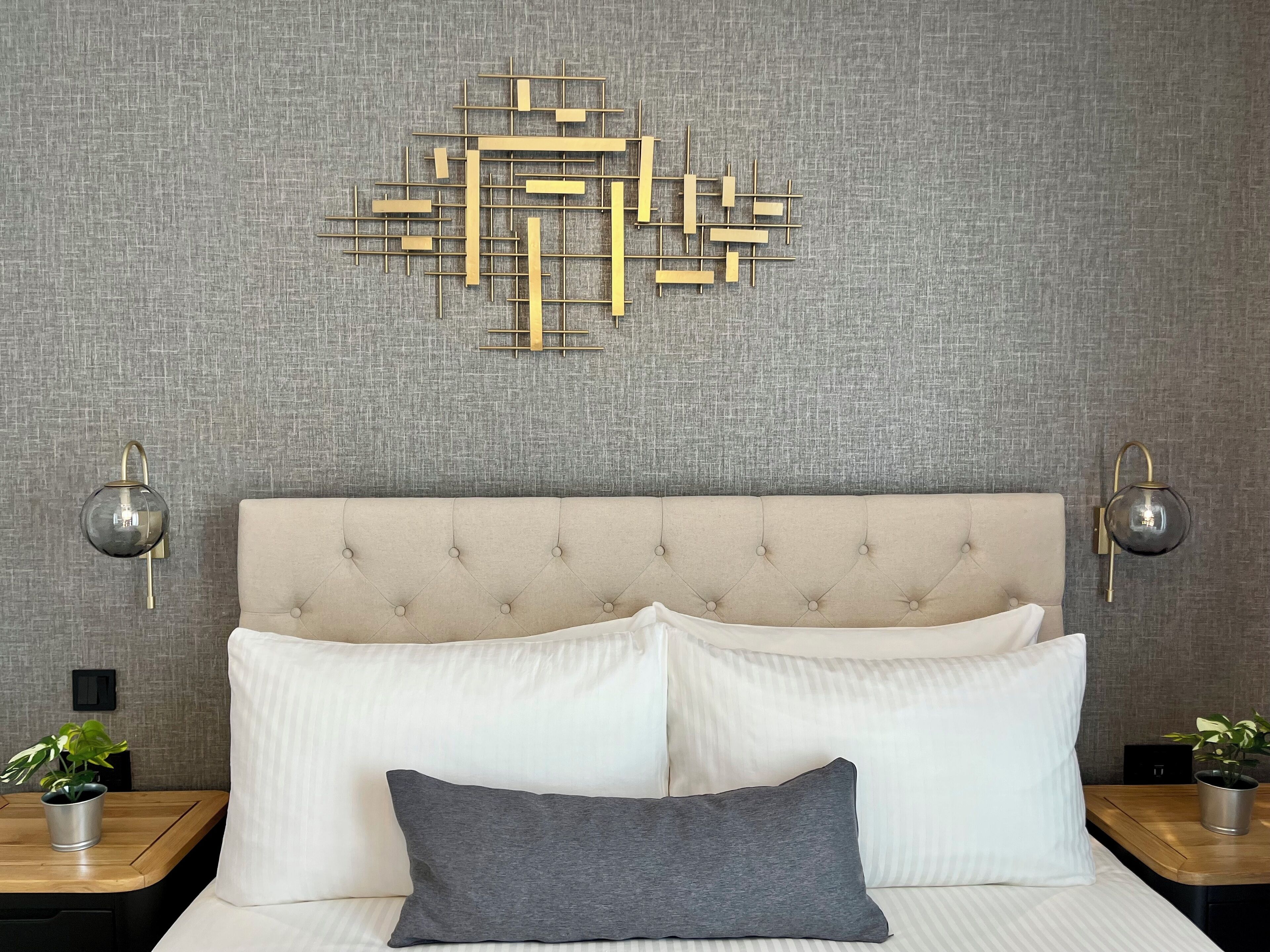 Una cama bien hecha con almohadas blancas y grises, un cabecero capitonado y una obra de arte geométrica en la pared.