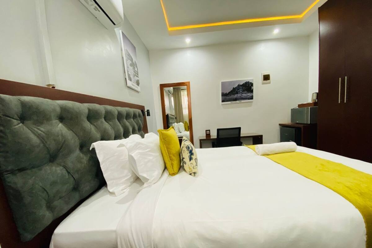 A hotel room with a large bed, a wooden headboard, a desk with a chair, a wardrobe, and a mirror on the wall.
