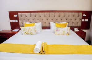 A neatly made bed with a yellow towel and two decorative pillows, a wooden headboard, and a bedside table with a phone and a notepad. - Bethel Suites (Bulawayo)
