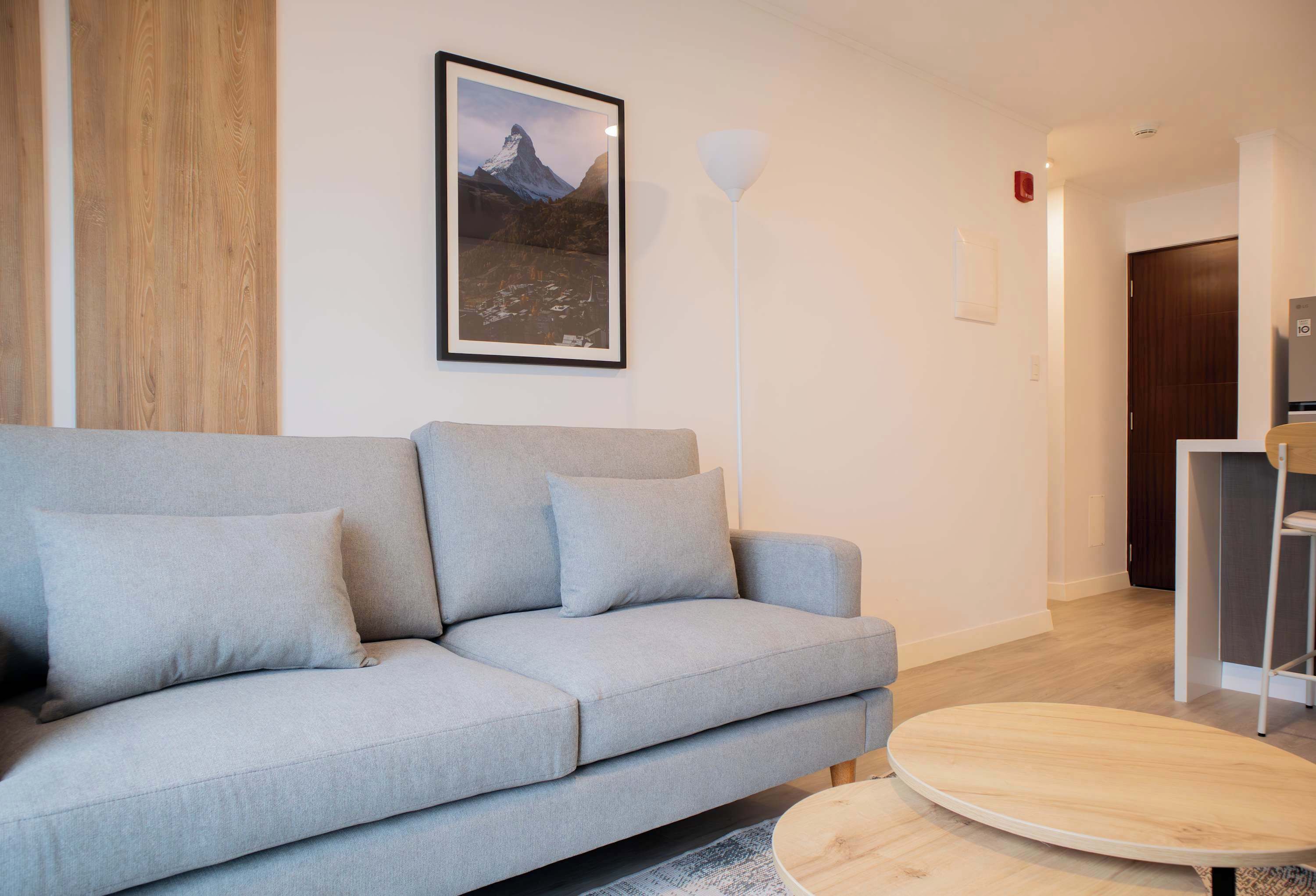 Ett modernt vardagsrum med en grå soffa, en inramad tavla med ett bergsmotiv och en vit golvlampa.