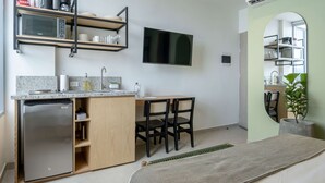 Una cocina compacta con refrigerador integrado, microondas y estantería. Hay un rincón de comedor con dos sillas y una mesita. Un televisor de pantalla plana está montado en la pared.