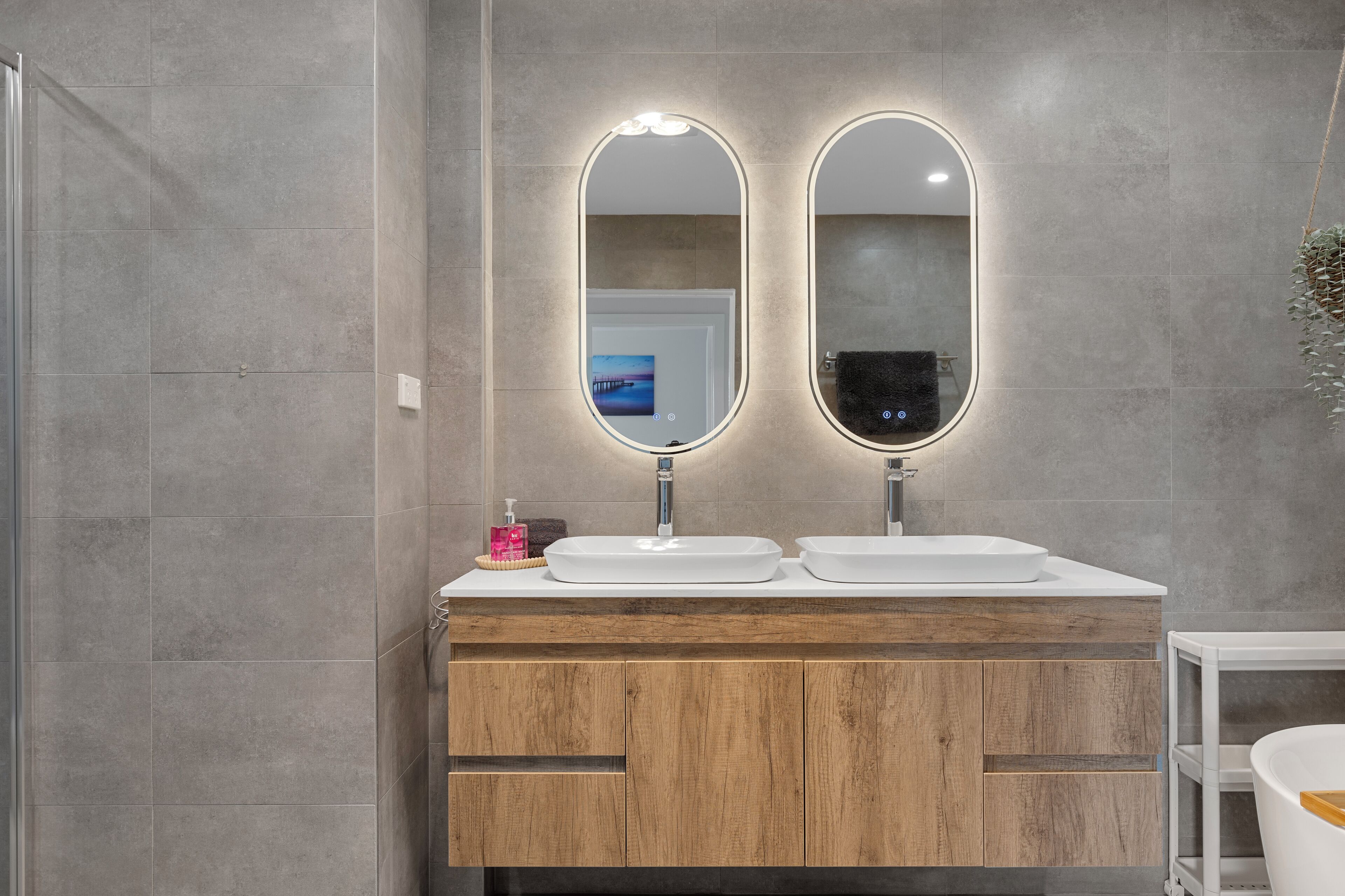 A modern bathroom with two oval mirrors, wooden vanity, and a shower area.