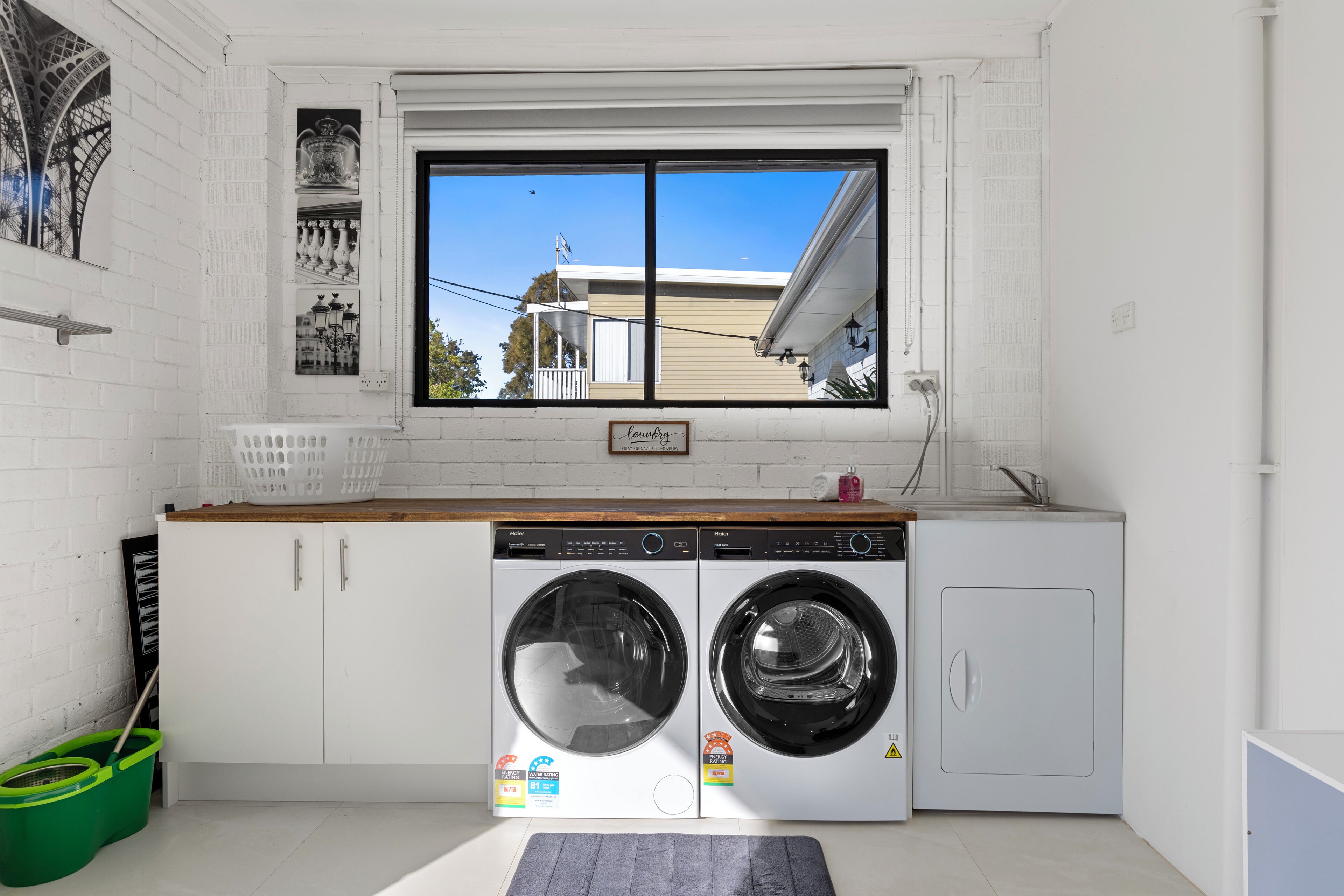 A laundry room with two front-loading washing machines, a window with a view of a house, and a wooden counter.