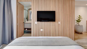 A modern bedroom with a large bed, a mounted TV, wooden panel walls, a potted plant, and a bathroom visible through an open door.