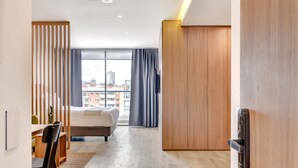 A modern hotel room with a bed, a desk, and a view of the cityscape through large windows.